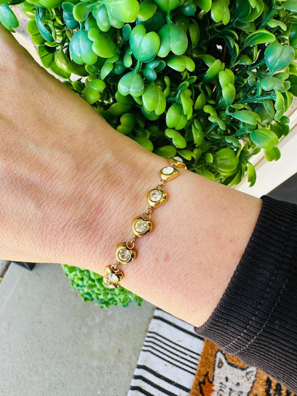 Gold-tone crystal heart bracelet worn on a wrist beside green leaves.
