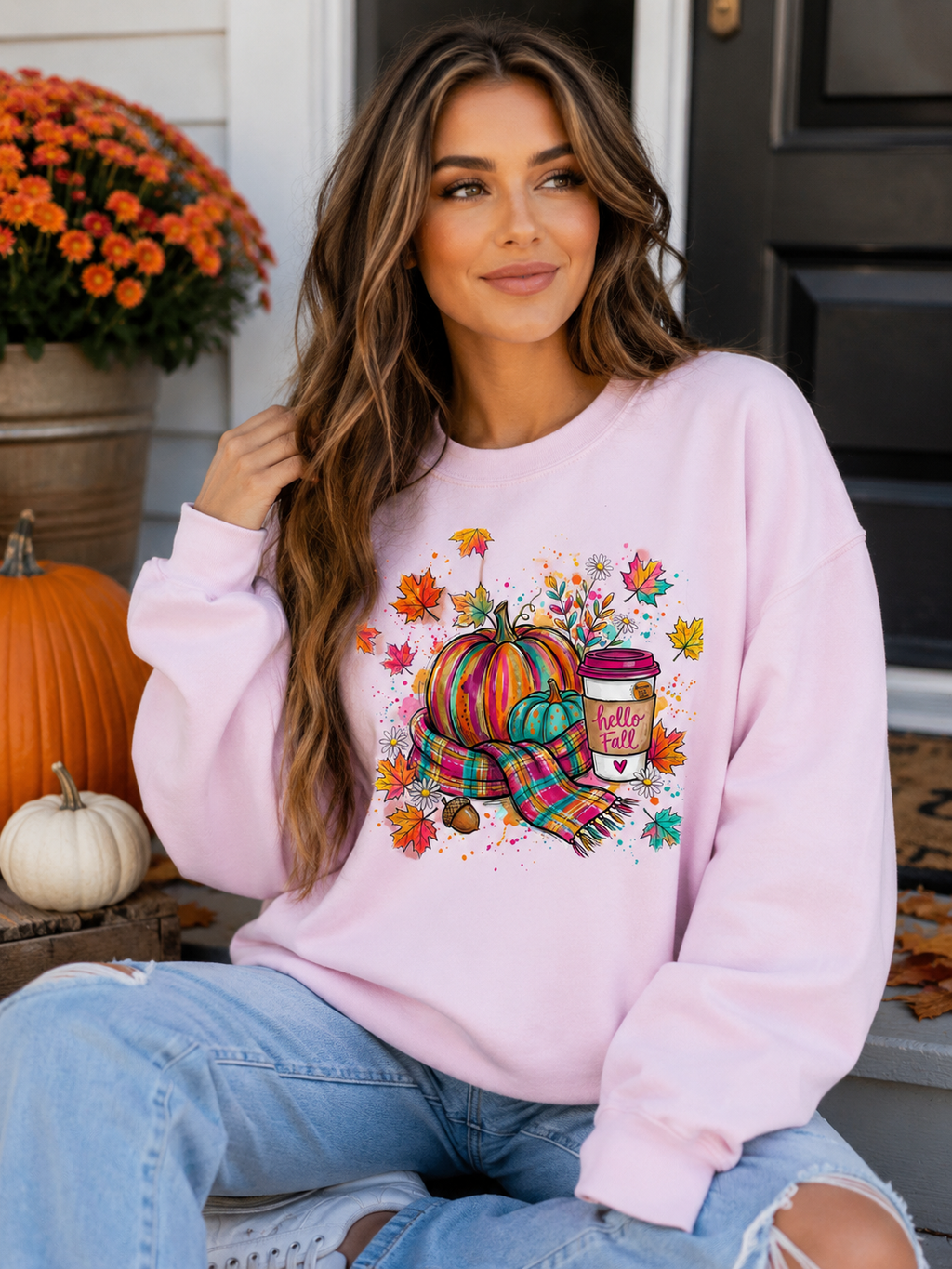 Woman wearing a white Hello Fall graphic sweatshirt with colorful pumpkins, leaves and a takeaway cup, styled with ripped jeans.