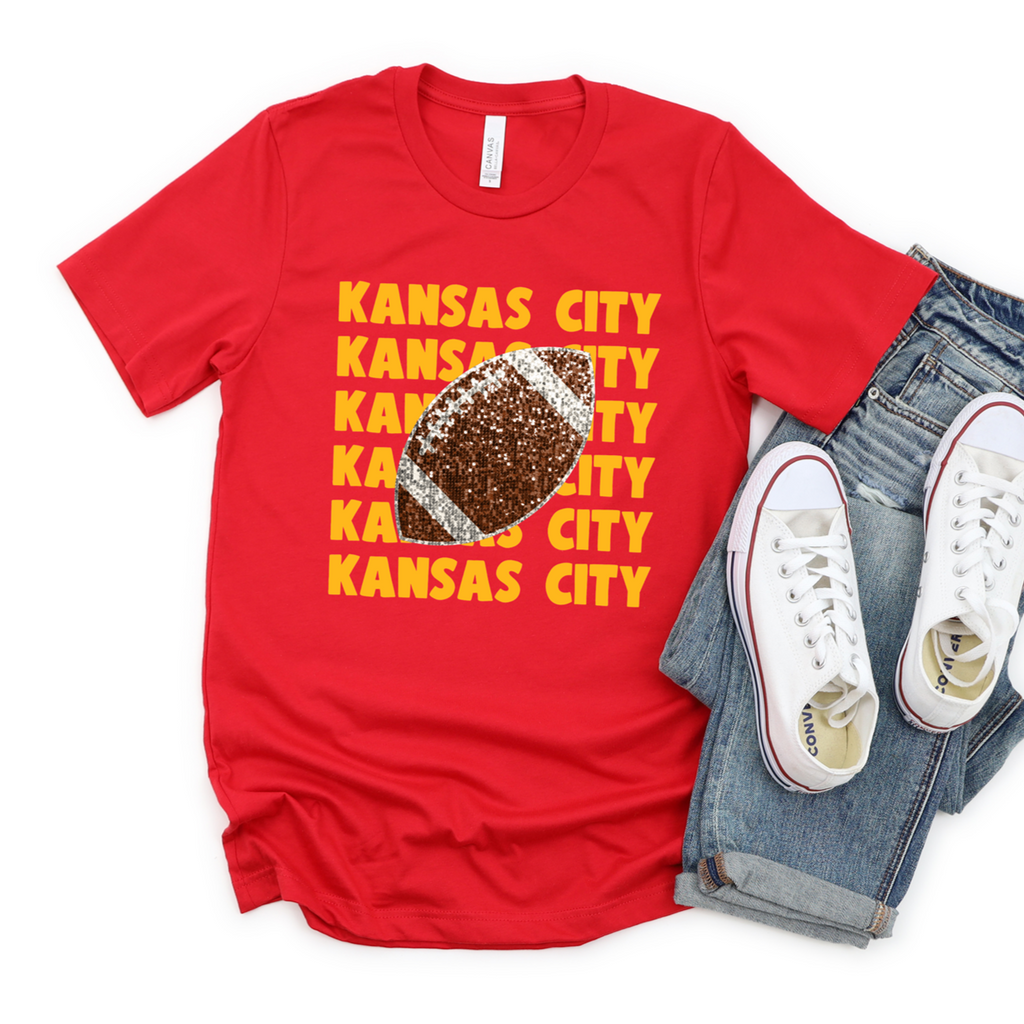 Model wearing a red crewneck with repeated yellow Kansas City lettering and a faux glitter football graphic.